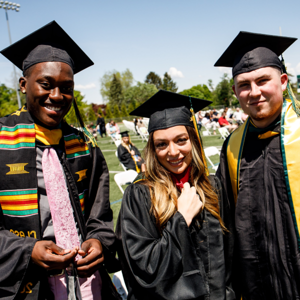 Three Students with cap and gown looking into 的 camera on 的 field at commencement. 